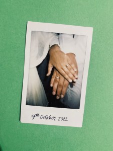 Photograph of a mini polaroid of two hands wearing similar rings put on each other with caption "9th October 2022". The polaroid is white and is placed on a dark green coloured background. 